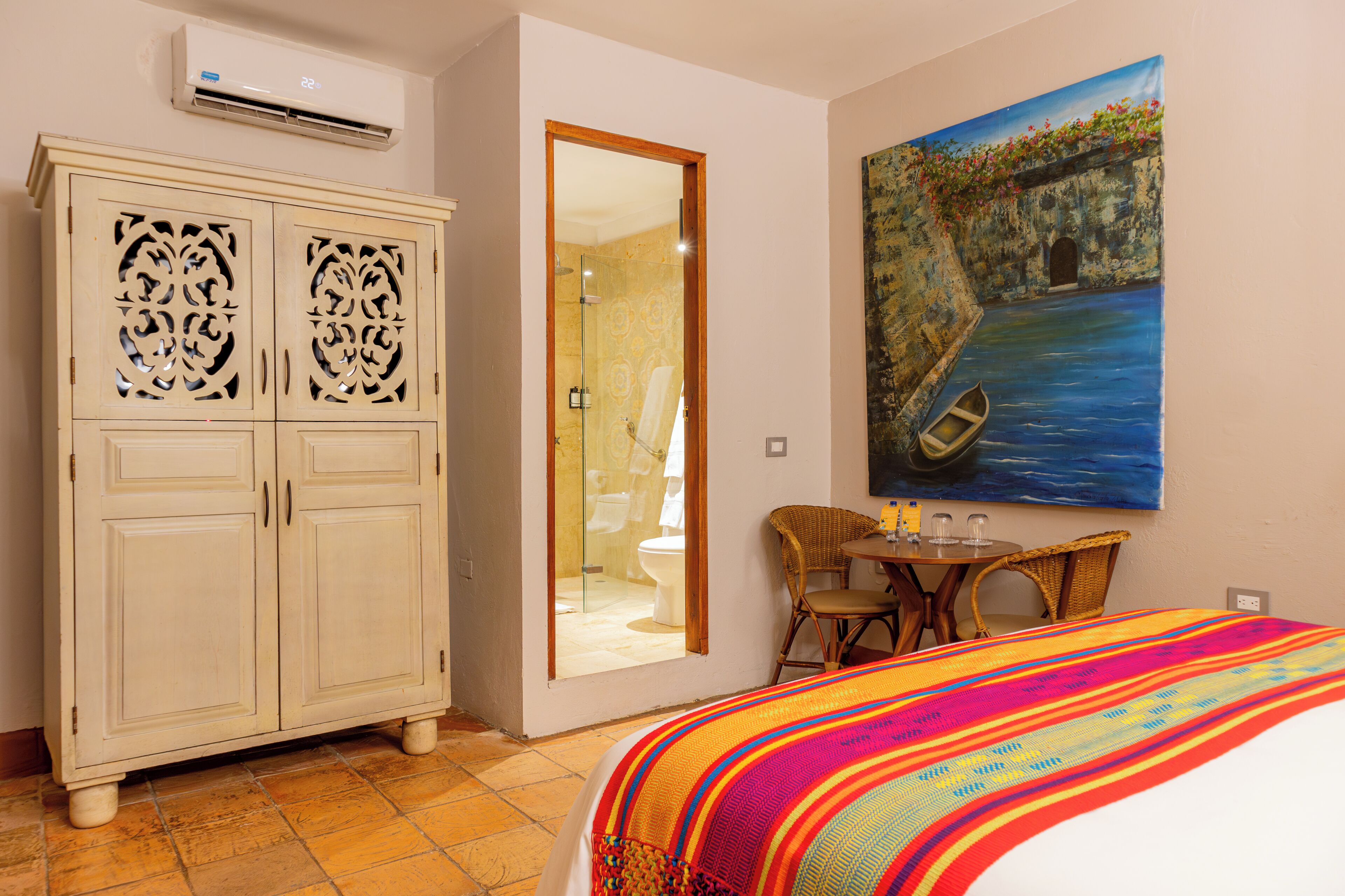 A hotel room with a bed, a wardrobe, a painting, a bathroom, and a small table with chairs.