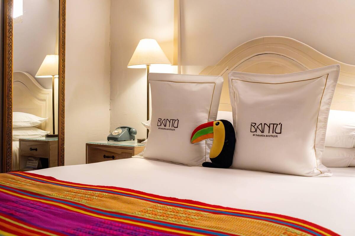 Una habitación de hotel con cama, almohadas, mesita de noche, lámpara y espejo.