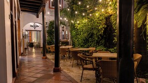 Un patio techado al aire libre con mobiliario de mimbre, luces colgantes y abundante vegetación.