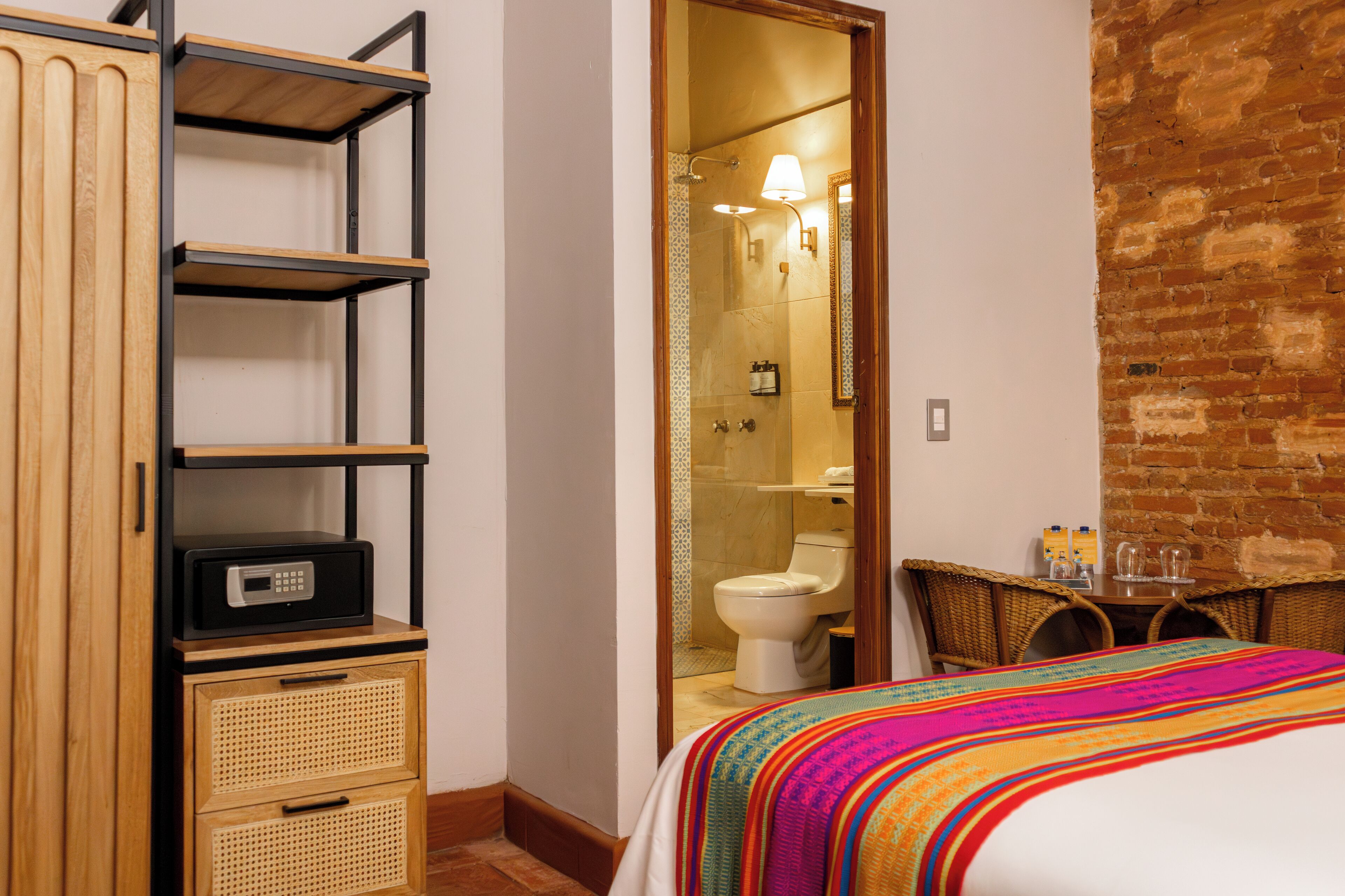 Una habitación de hotel con un armario de madera, una cama con un edredón colorido, una mesita con vasos y una pared de ladrillo.