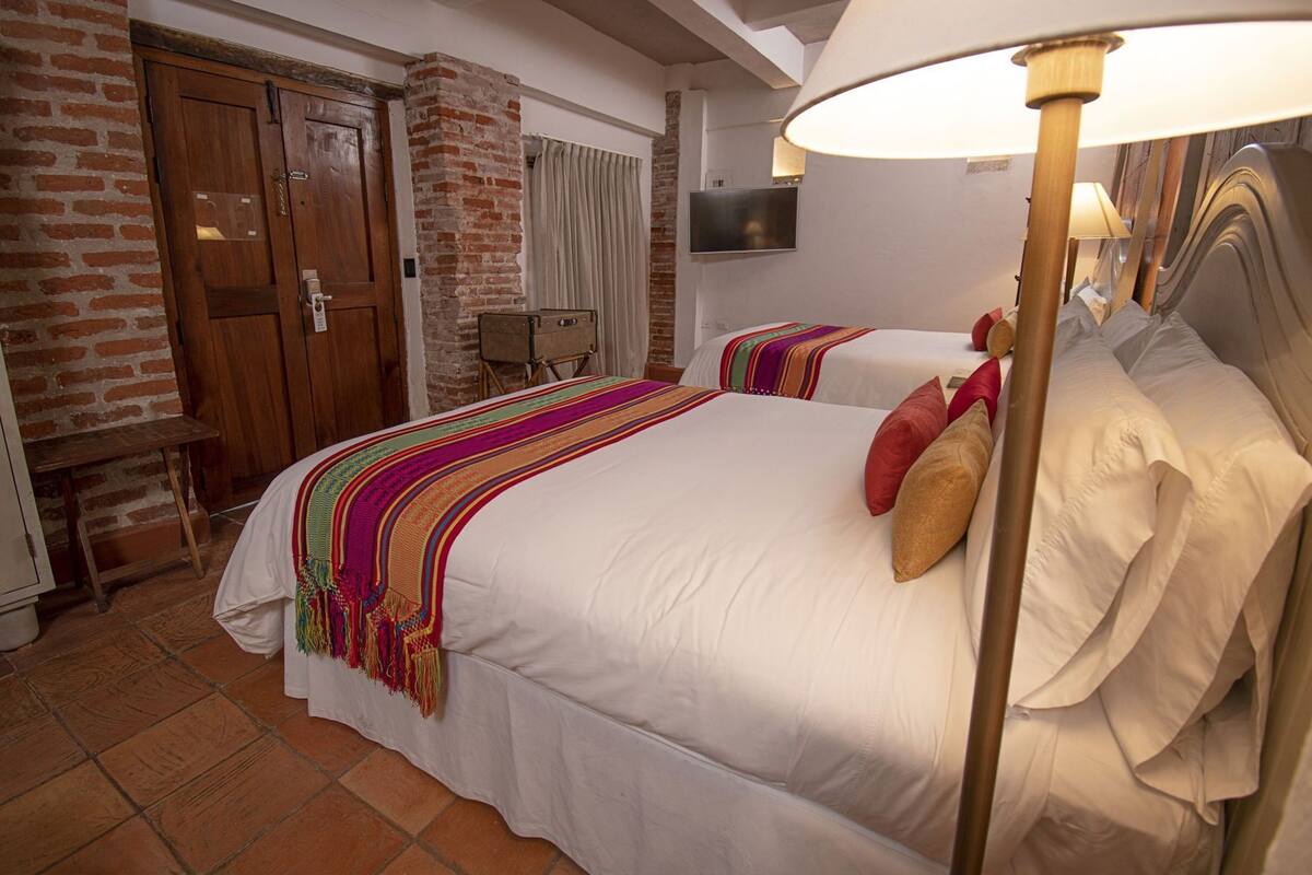 Habitación de hotel con dos camas, una pared de ladrillo, una puerta de madera, un televisor y una lámpara.