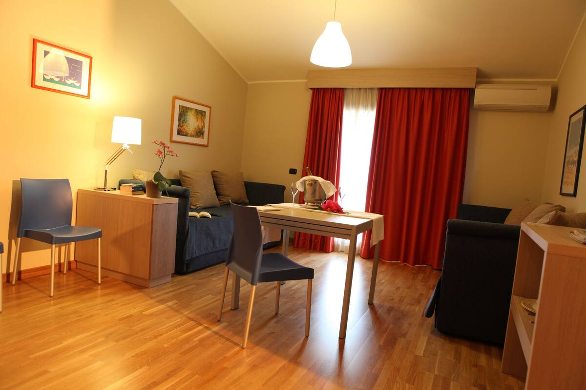 A modern living room with a dining area, wooden flooring, and red curtains.