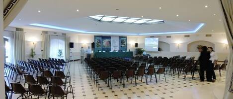 A conference room with rows of chairs, a stage with a green backdrop, and a high ceiling with skylights.