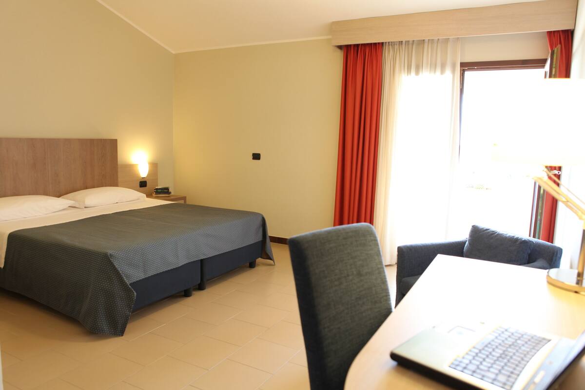 A hotel room with a bed, a desk with a laptop, a chair, and a window with curtains.