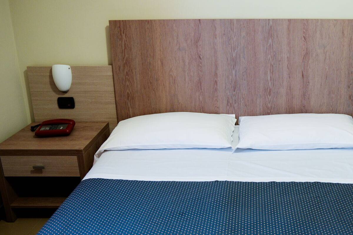 A neatly made bed with white linens and a blue patterned bedspread, flanked by wooden nightstands with a red telephone and a black object on one, and two wall-mounted light fixtures on the other.