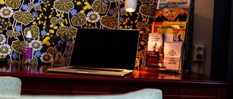 A laptop on a desk with a floral wallpaper, a lamp, and some magazines.