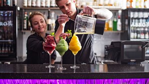 Un cantinero sirve una bebida en un vaso con una jigger. Hay tres bebidas coloridas sobre la barra.