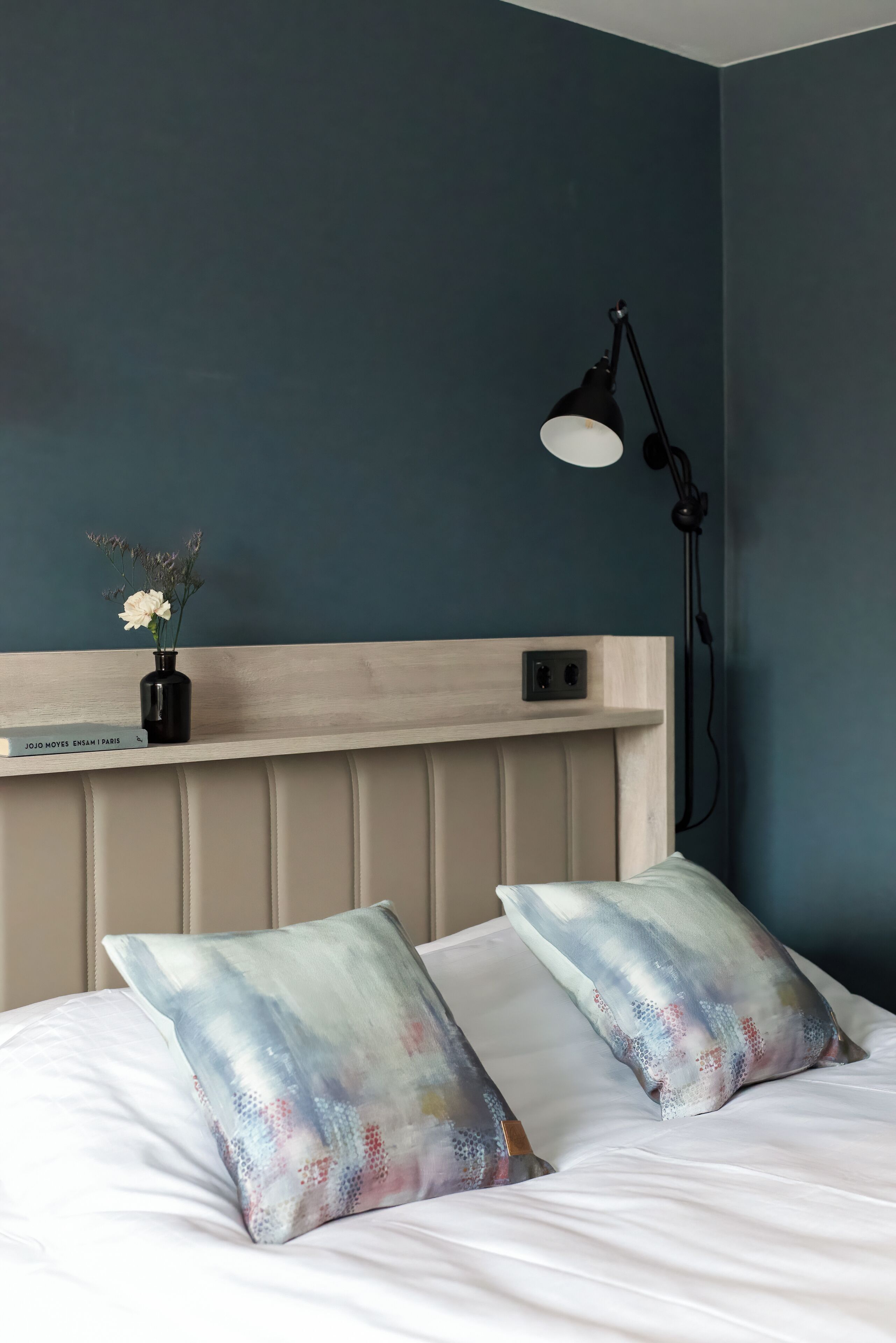 A bedroom with a bed, two pillows, a nightstand, a lamp, and a vase with flowers.