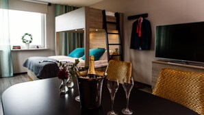 Habitación de hotel con literas, un televisor, una mesa de comedor con una botella de champán y copas, y una ventana con cortinas.