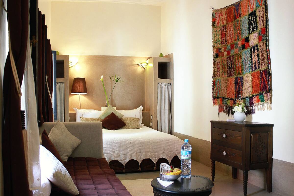 A hotel room with a bed, a sofa, a small table with a bottle and fruit, a tapestry, and an air conditioning unit.