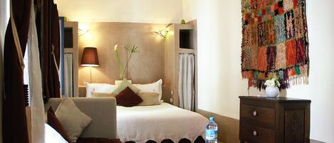 Ein Hotelzimmer mit Bett, Sofa, kleinem Tisch mit Flasche und Obst, Wandteppich und Klimaanlage.