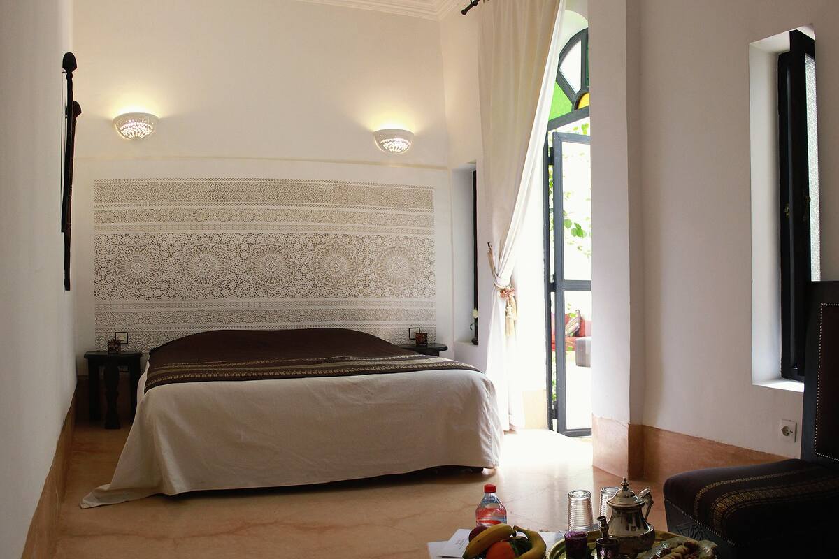 A bedroom with a bed, a nightstand, a table with fruits and drinks, and a view of the outside.