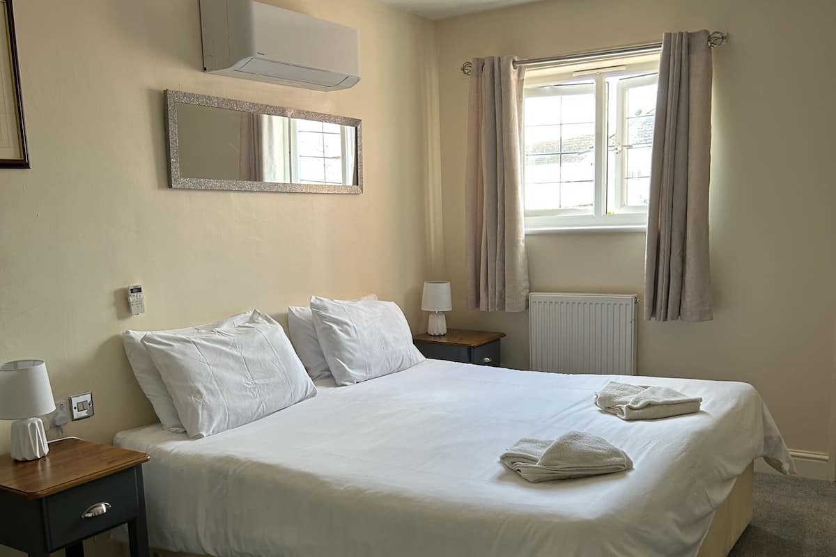 A neatly made bed in a hotel room with a bedside table, a lamp, and a window with curtains.