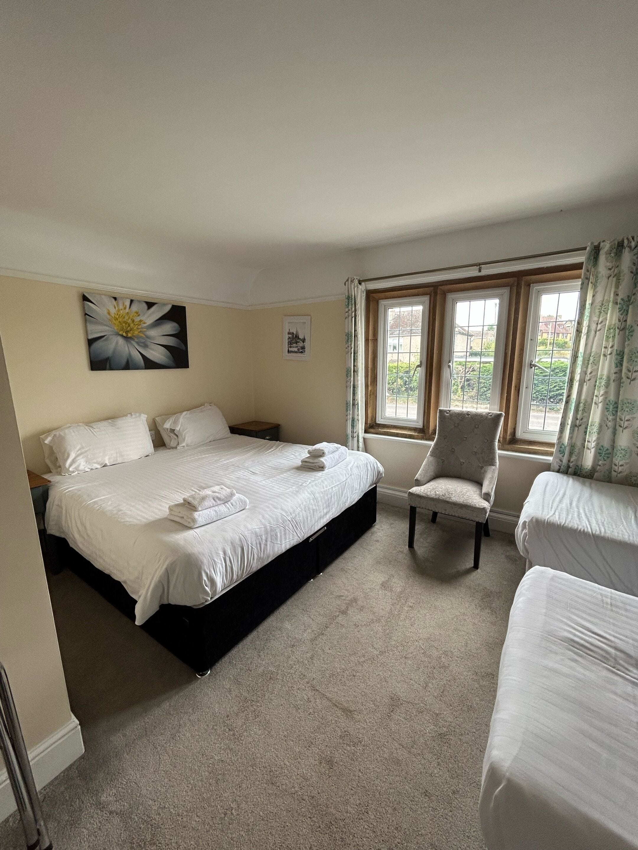 Quarto de hotel com uma cama grande, uma cadeira, janela com cortinas e um quadro de flores na parede.