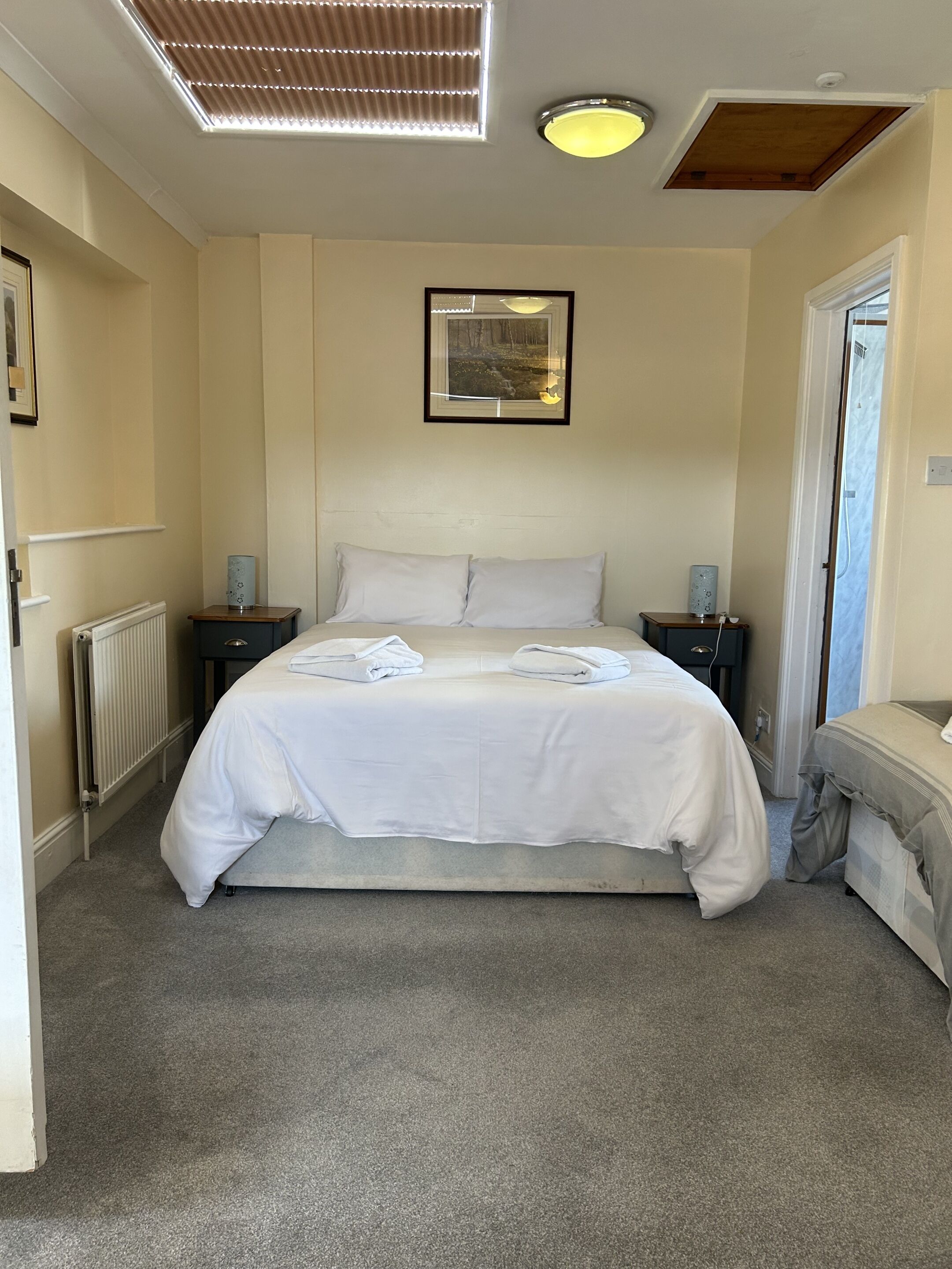 Une chambre avec un grand lit, deux tables de chevet, un radiateur, un luminaire au plafond, un tableau encadré et une porte donnant sur une autre pièce.