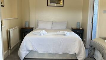 A bedroom with a large bed, two bedside tables, a radiator, a ceiling light, a framed picture, and a door leading to another room.