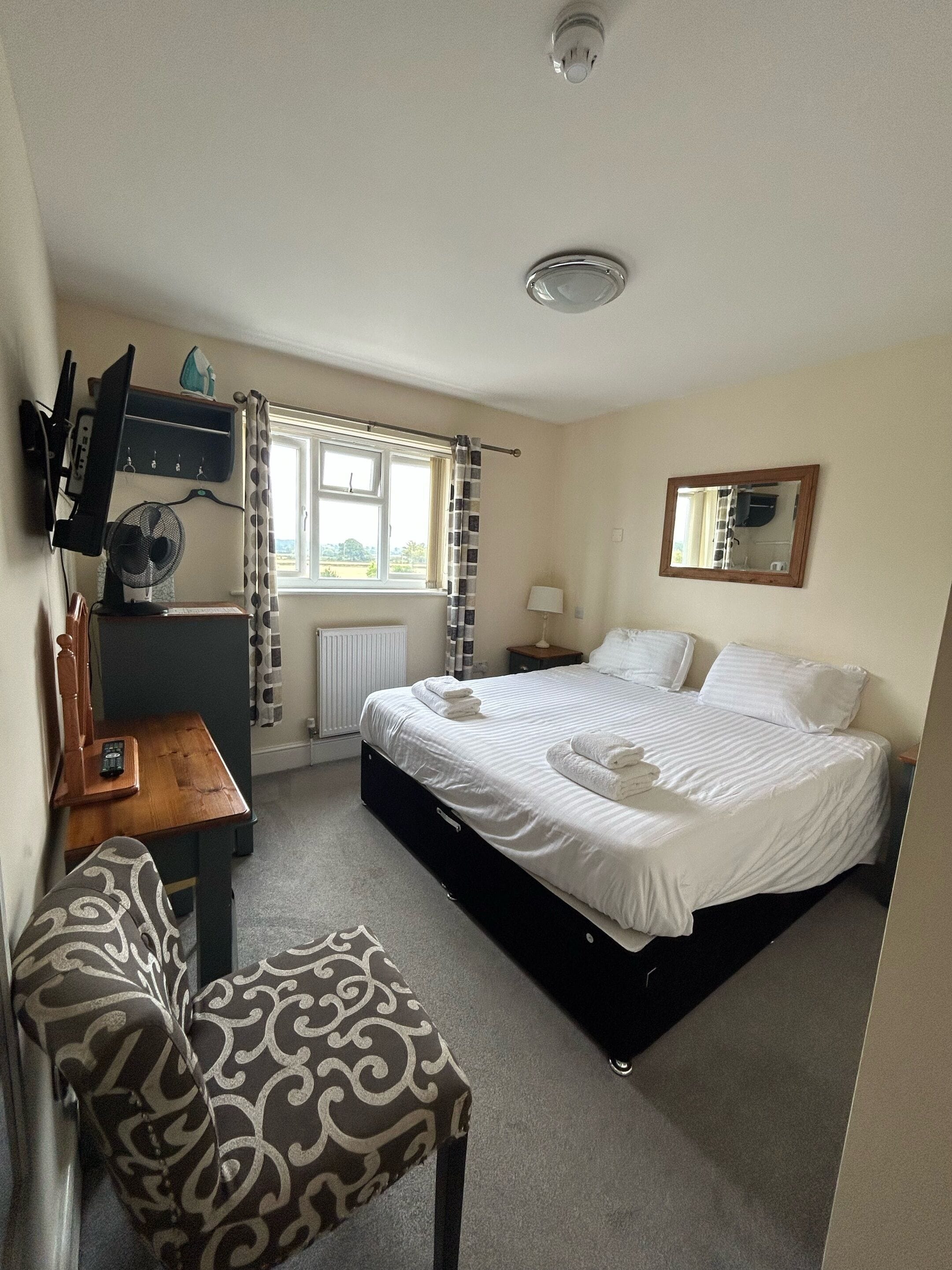 A bedroom with a bed, a desk, a chair, a window with curtains, a bedside table, and a mirror on the wall.