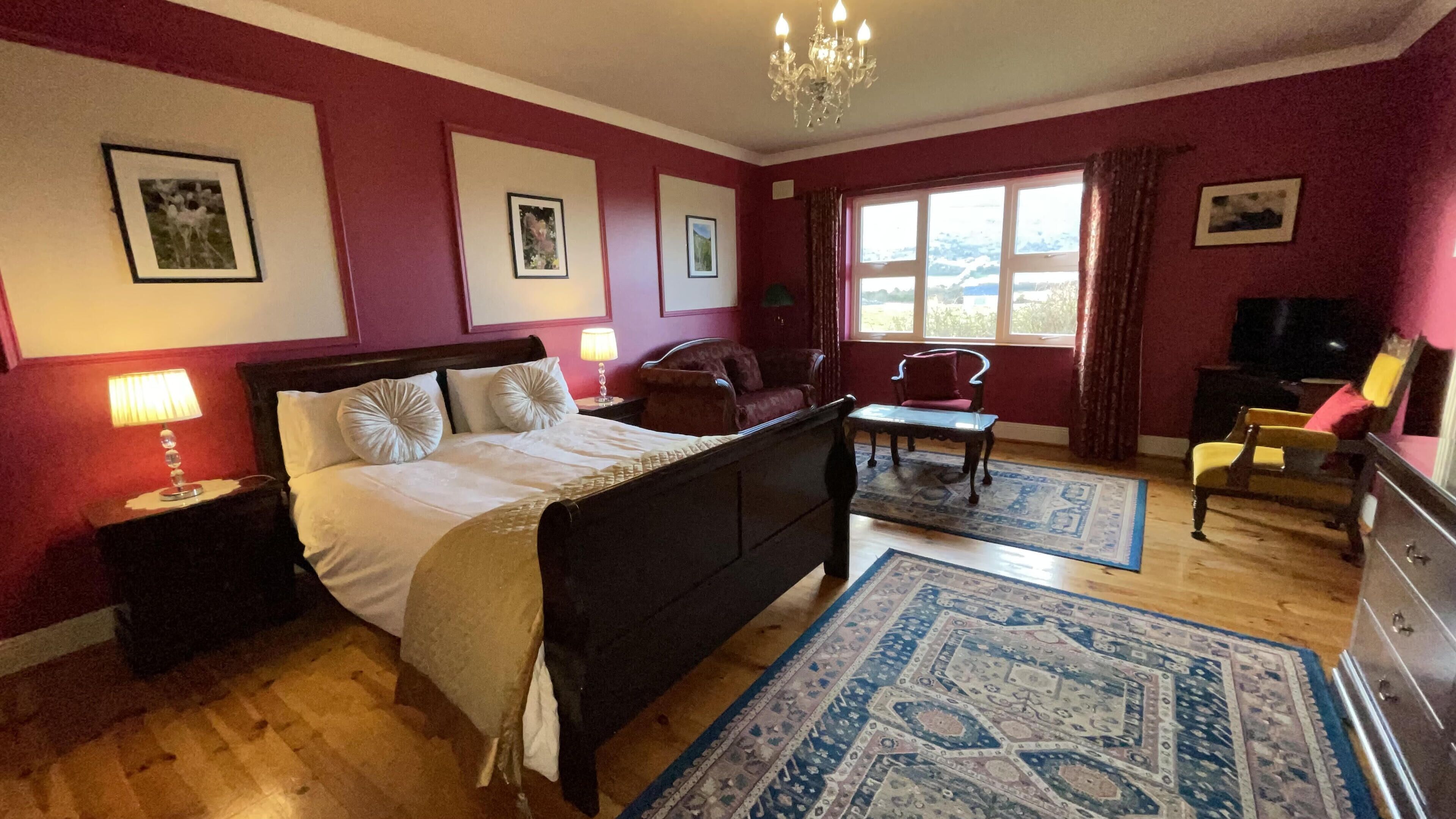 A bedroom with a bed, two chairs, a desk, a TV, and a chandelier.