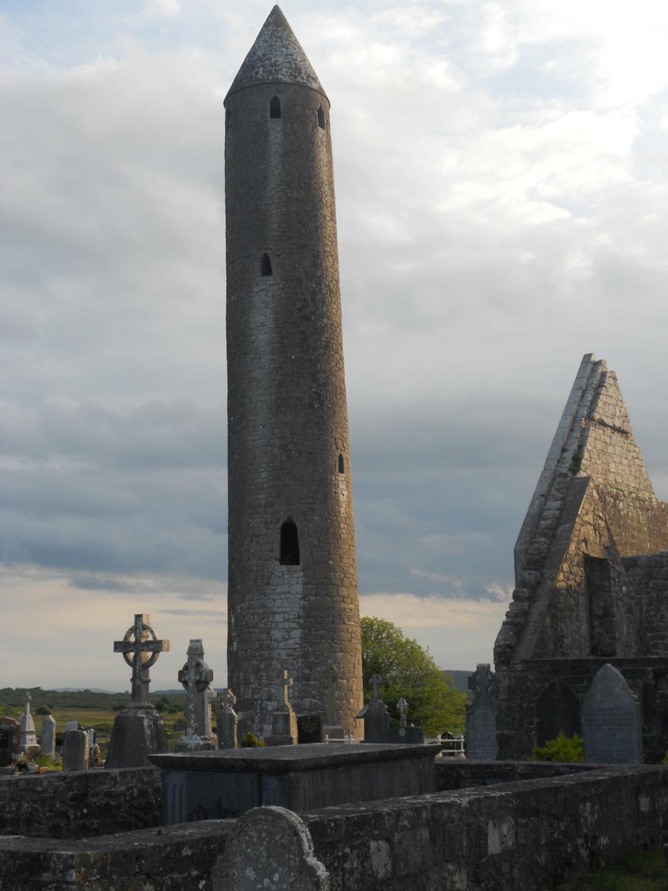 A tall, narrow stone tower with a pointed roof, surrounded by a cemetery with various gravestones and crosses.