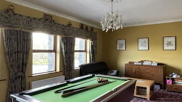 Una mesa de billar en una habitación con alfombra floral, un candelabro y una ventana con cortinas.