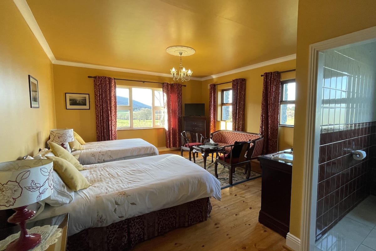 A hotel room with two beds, a dining table, a sofa, a chandelier, and a bathroom with a toilet and sink.
