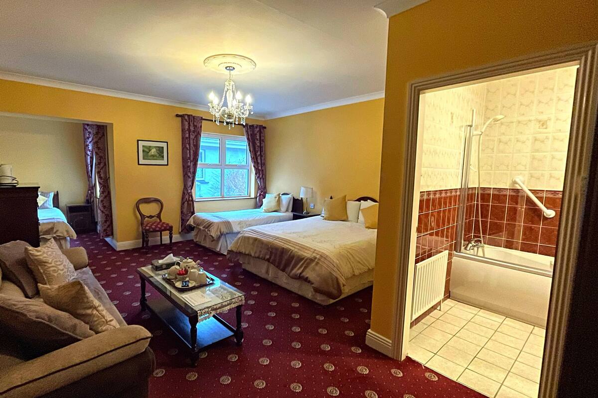 A hotel room with two beds, a sofa, a small table, a chandelier, and a bathroom visible through an open door.