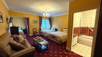 A hotel room with two beds, a sofa, a small table, a chandelier, and a bathroom visible through an open door.