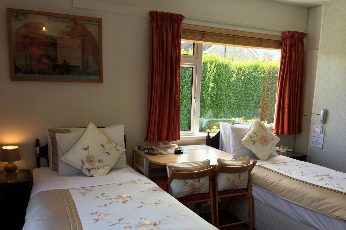 A hotel room with a bed, a desk, and a chair. There is a window with curtains, a painting on the wall, and a sign on the wall.