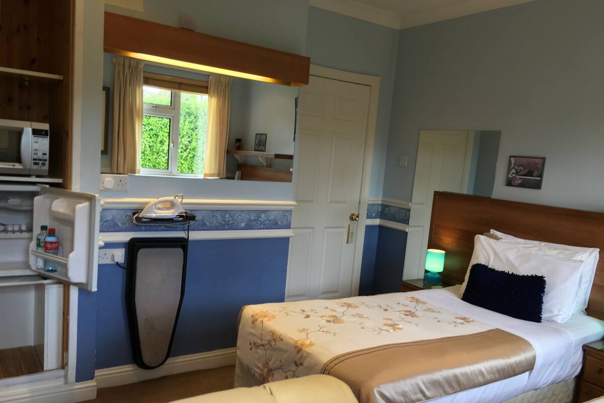 A hotel room with a bed, bedside table, lamp, and a small kitchenette area.