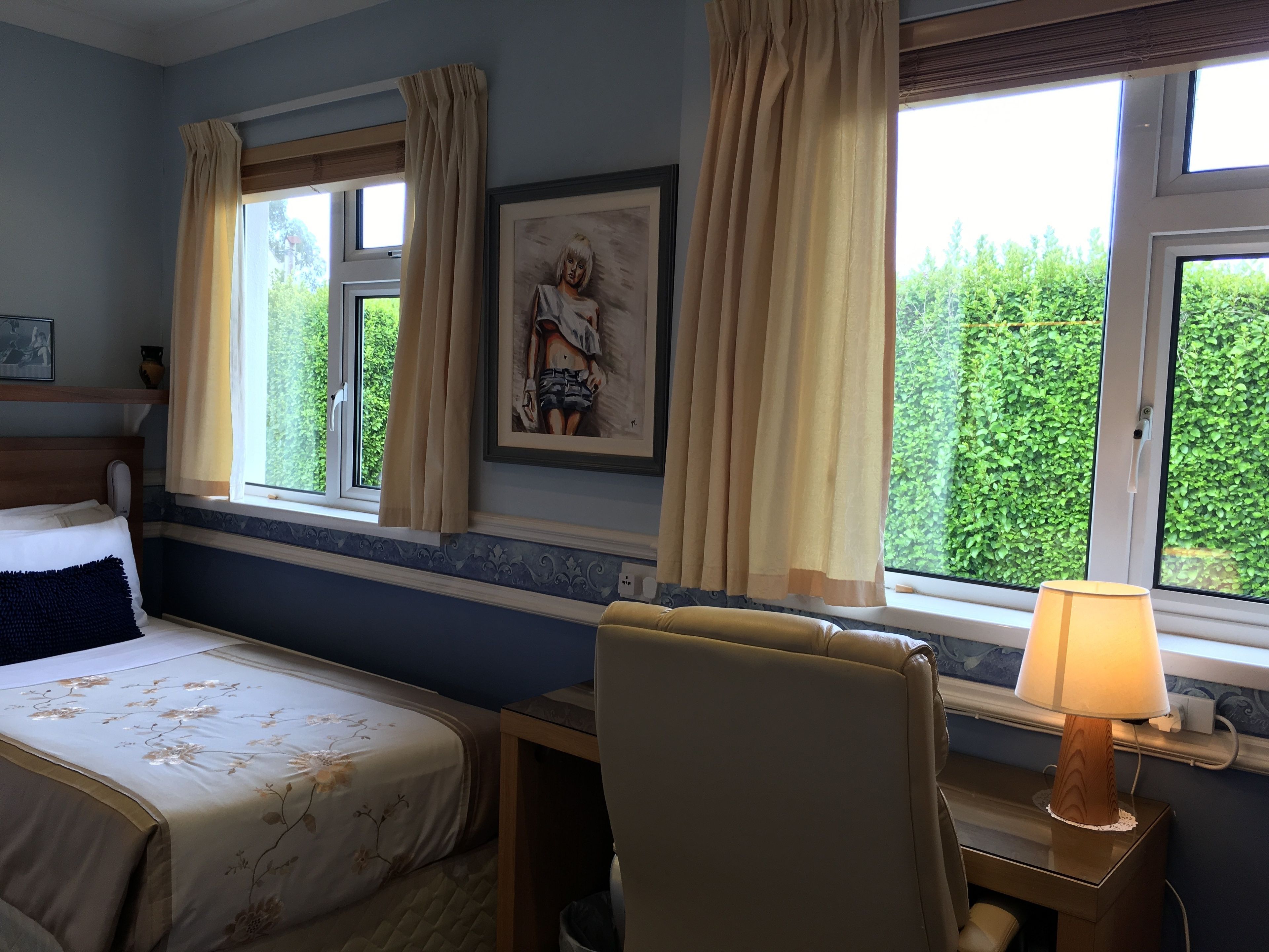 A hotel room with a bed, a desk with a lamp, a chair, a window with curtains, and a framed picture on the wall.