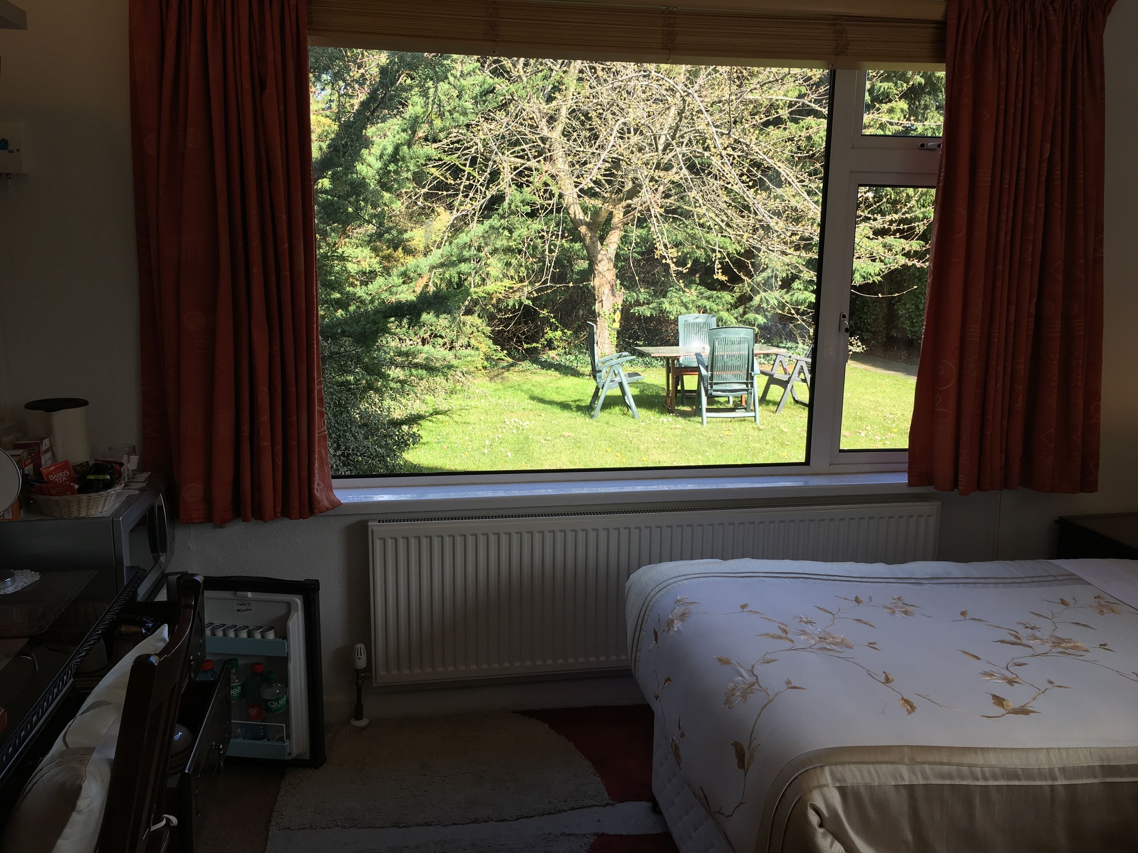 Un dormitorio con un ventanal grande, una cama, un radiador, un escritorio con lámpara, y vistas a un jardín con mesa y sillas.