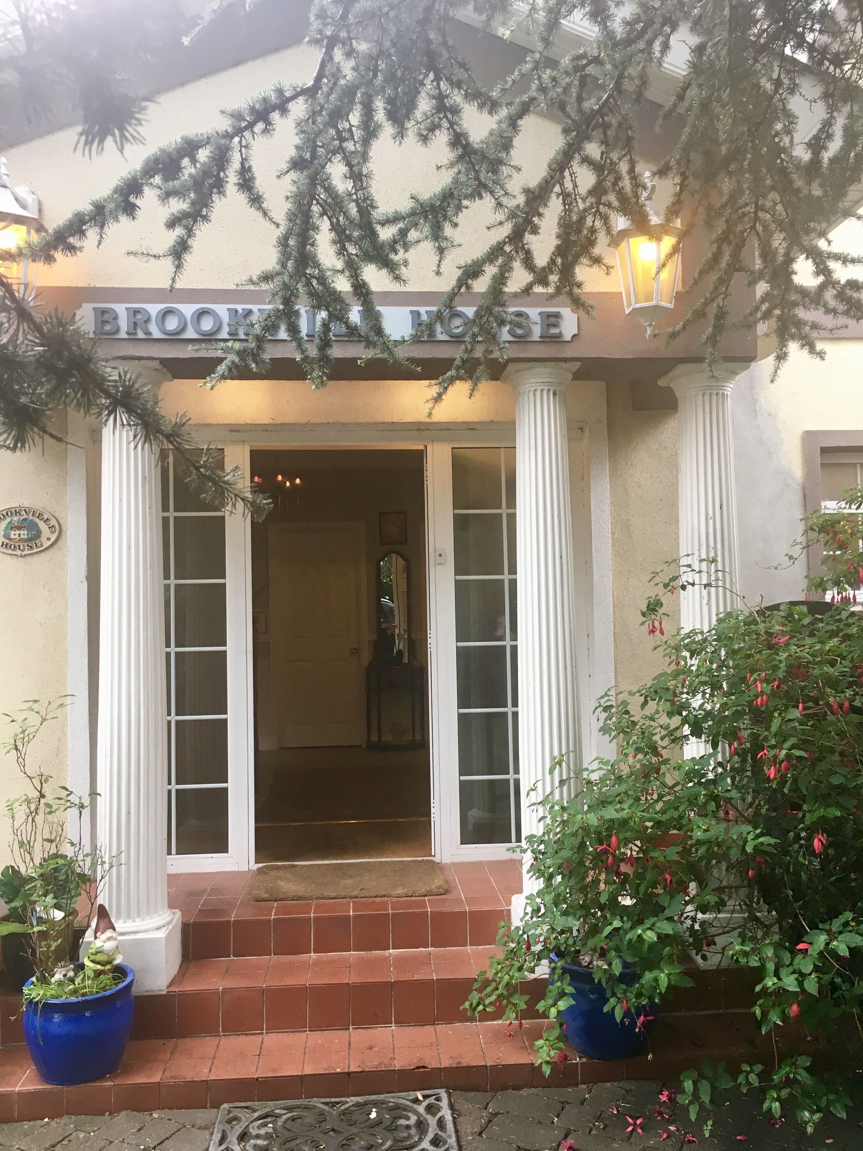 Η είσοδος του Brookhill House, με μια επιγραφή πάνω από την πόρτα, μια σειρά σκαλοπατιών που οδηγούν προς την είσοδο και δύο γλάστρες με φυτά εκατέρωθεν.