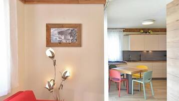 Ein modernes Wohnzimmer mit einem roten Sofa, einer Wandlampe und einem gerahmten Bild. Daneben befindet sich eine Küche mit einer Essecke und farbenfrohen Stühlen.