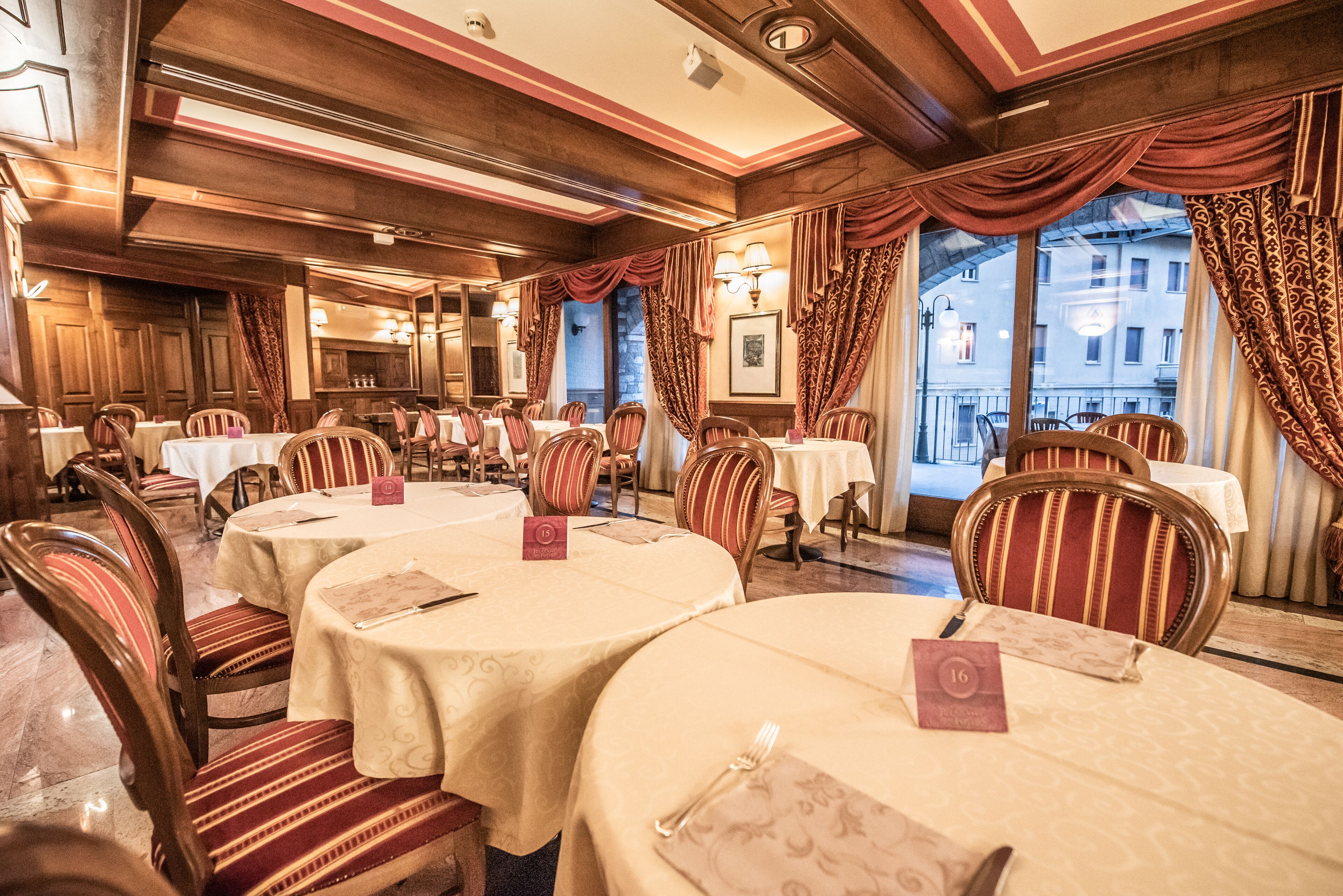 Un restaurante con mesas redondas, sillas a rayas y vistas a edificios a través de amplios ventanales.