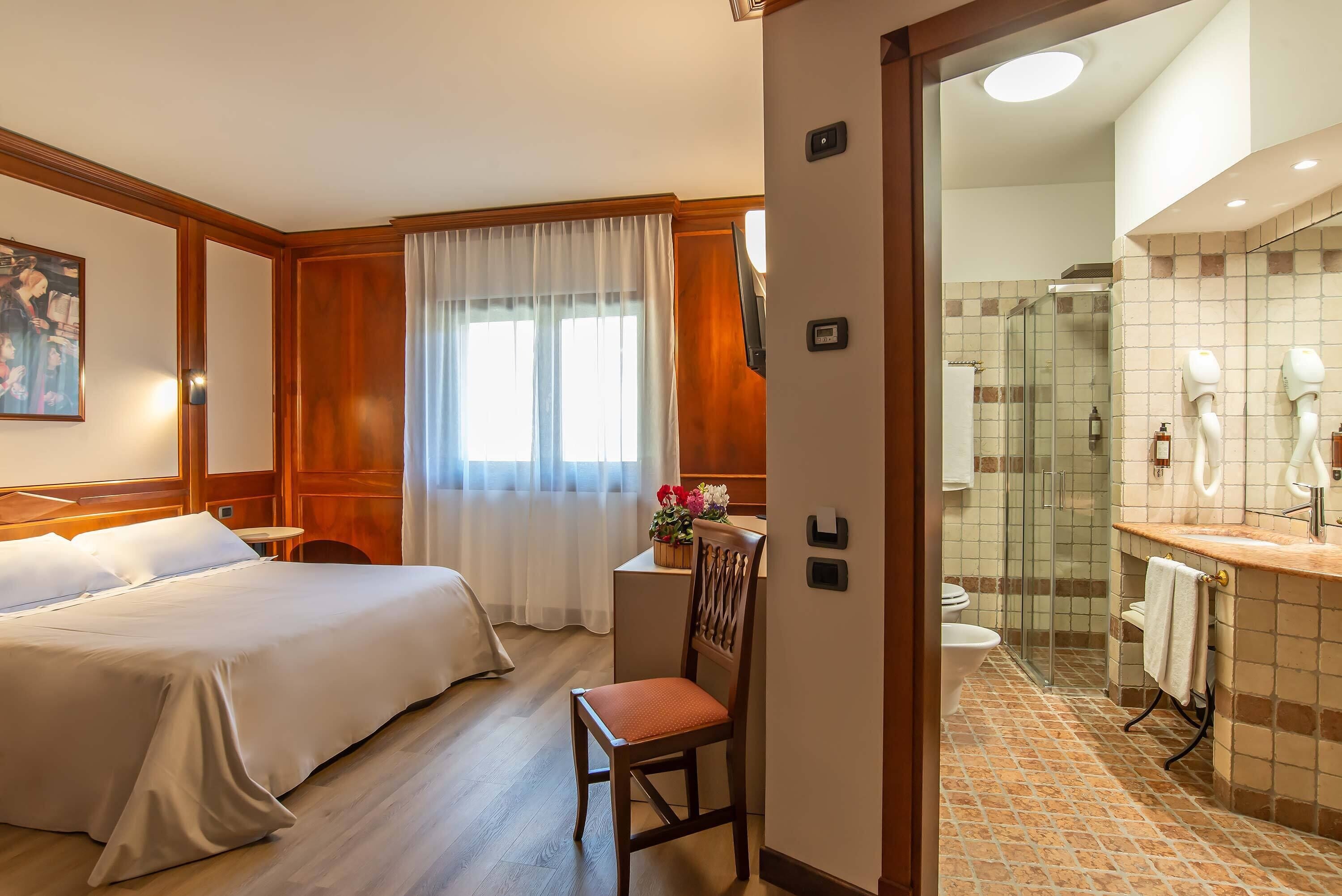 Habitación de hotel con cama, una silla, escritorio, ventana con cortinas, baño con ducha y lavabo.