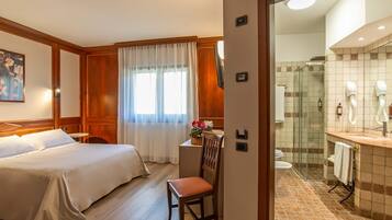 Una camera d'albergo con un letto, una sedia, una scrivania, una finestra con tende, un bagno con doccia e un lavandino.