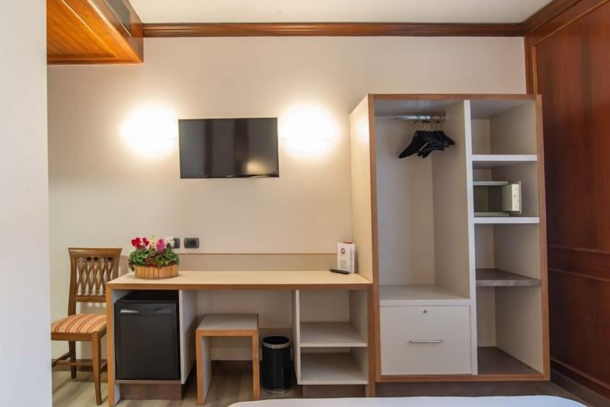 Habitación compacta con cama, escritorio con silla, refrigerador pequeño, televisión montada en la pared y armario con estantes.
