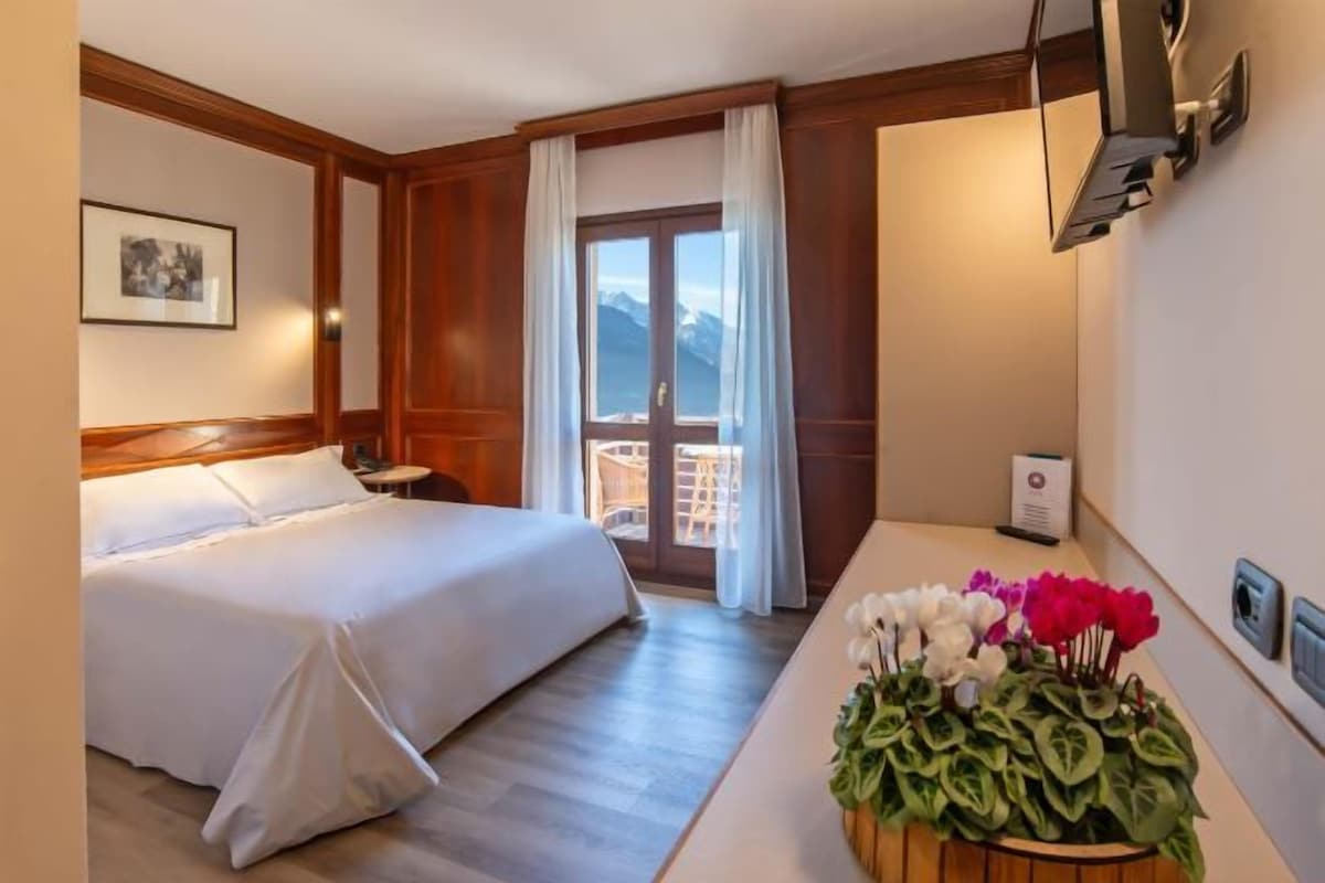 A hotel room with a bed, a small table with a plant, a window with curtains, and a door leading to a balcony.