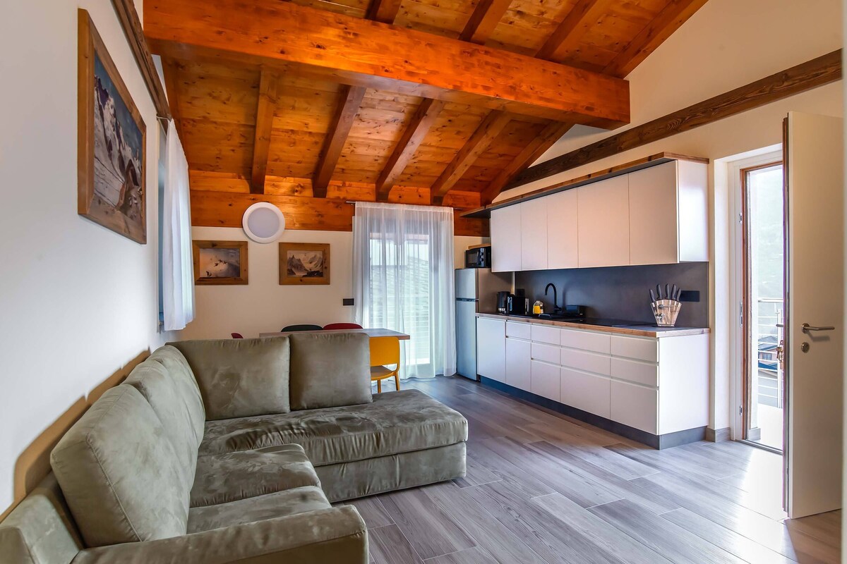 Una sala de estar con un sofá, vigas de madera en el techo, una cocina con armarios blancos y una zona de comedor con una mesa y sillas.