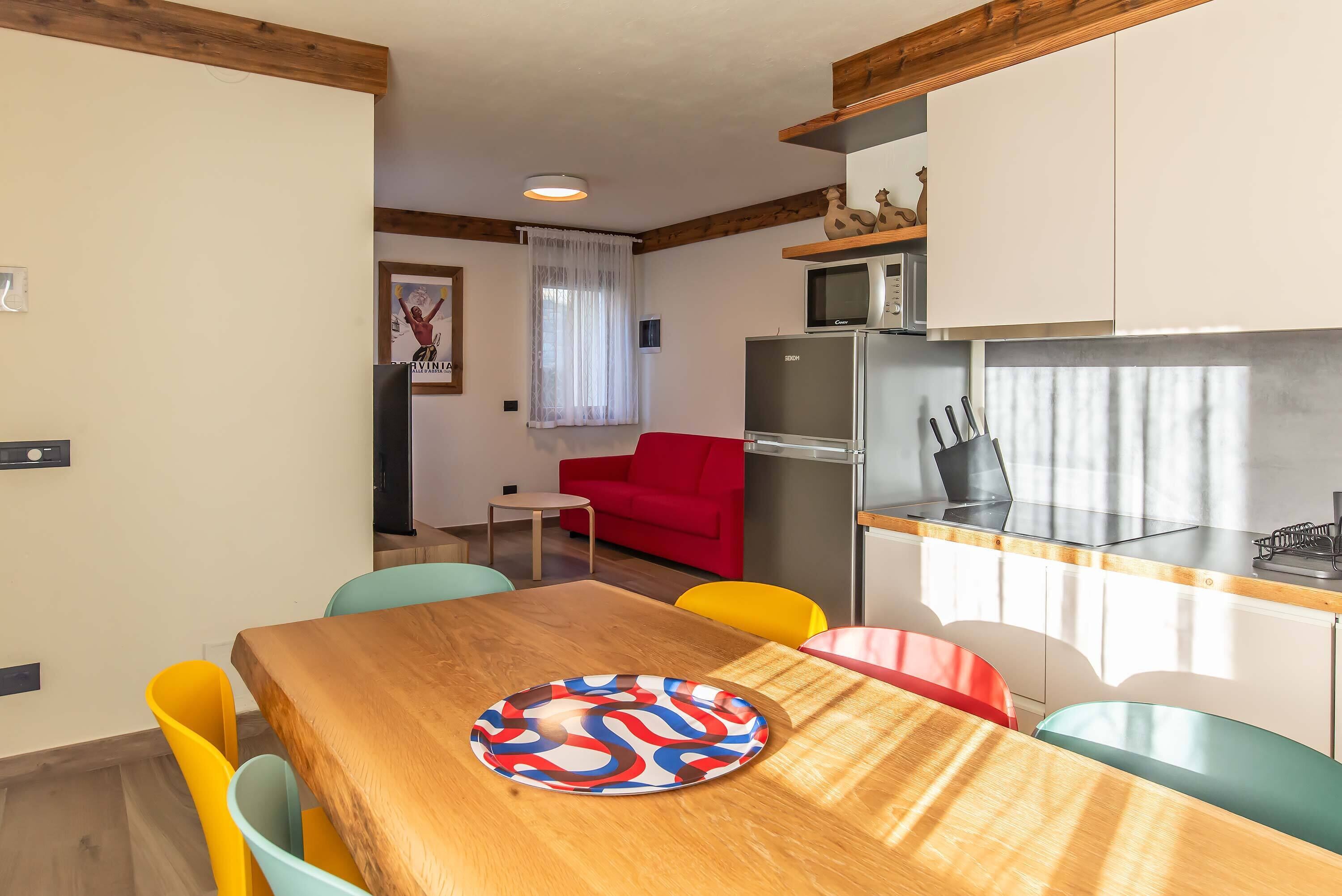Un espace de vie compact comprenant une table à manger en bois, des chaises colorées, un canapé rouge, un coin cuisine avec un four à micro-ondes et un réfrigérateur, ainsi qu’une petite fenêtre avec des rideaux.