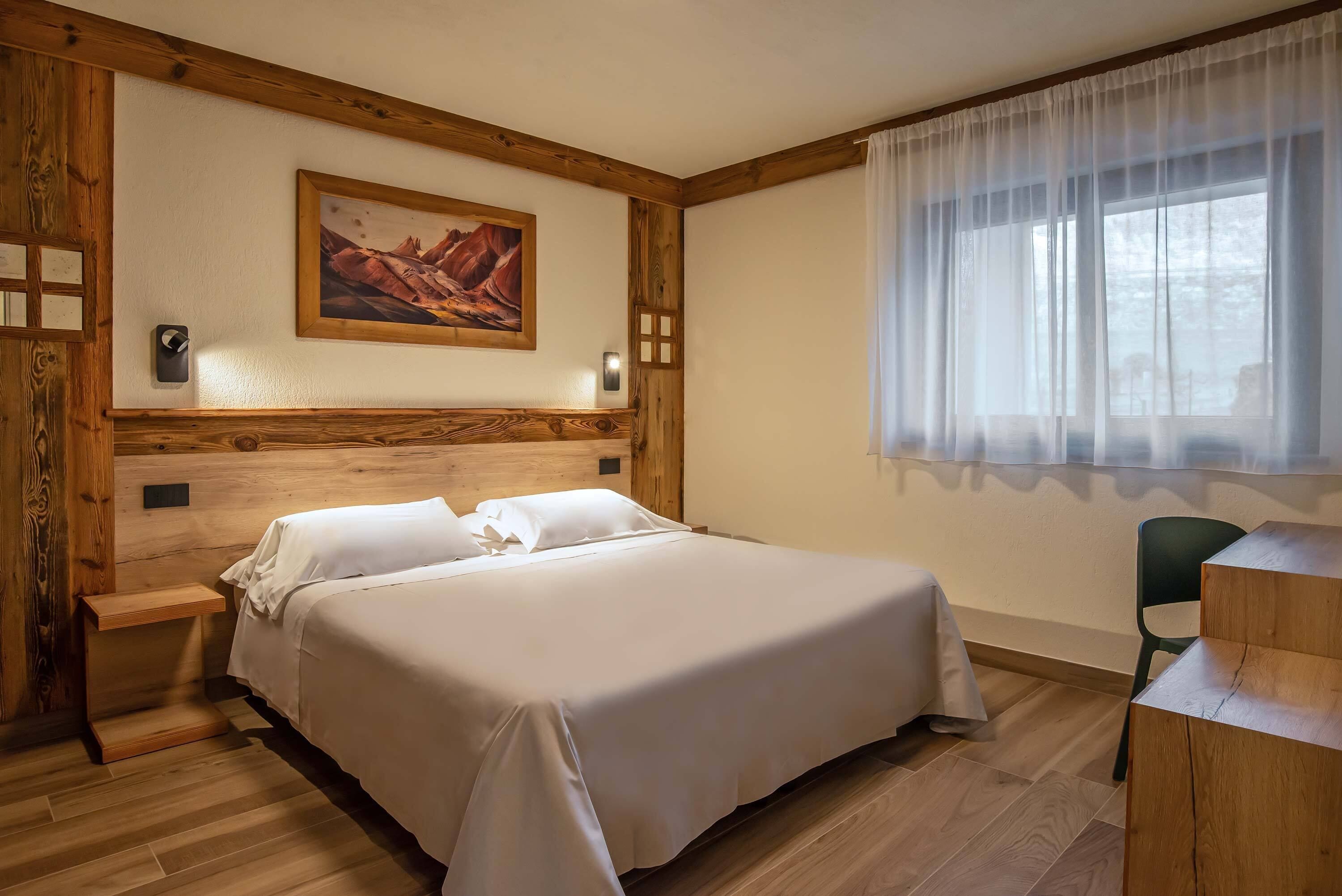 Un dormitorio ordenado con tablero de cama de madera, ropa de cama blanca, mesita de noche, escritorio y una ventana grande con cortinas traslúcidas.