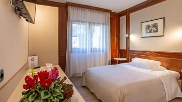 Ein Hotelzimmer mit einem Bett, einem Nachttisch mit Blumen, einem Fenster mit Vorhängen, einem an der Wand montierten Fernseher und einem gerahmten Bild.