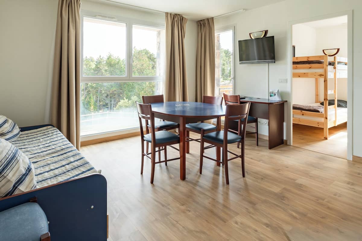 A room with a wooden floor, a dining table with chairs, a sofa, a TV mounted on the wall, and a bunk bed.