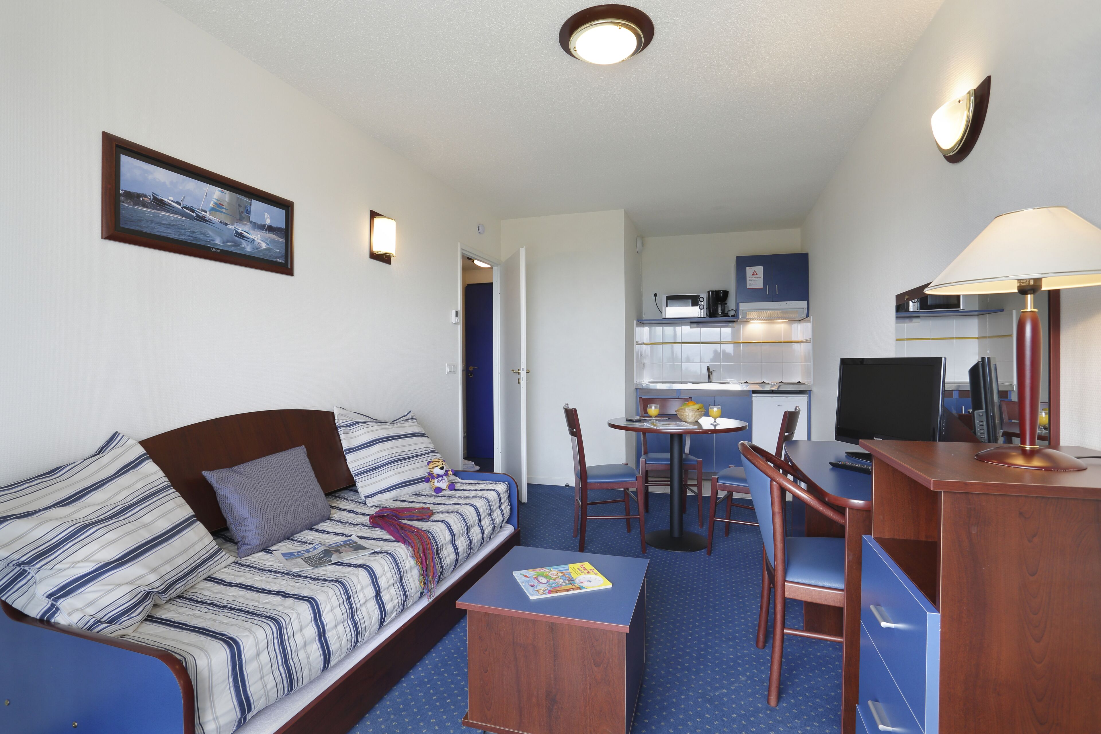 Ett kompakt hotellrum med en enkel säng, ett litet skrivbord med en lampa, en tv och ett litet pentry i bakgrunden.