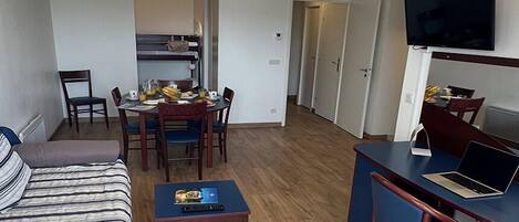 Habitación de hotel compacta con sofá, una pequeña mesa de comedor con sillas, kitchenette, televisor montado en la pared y un escritorio con ordenador portátil.