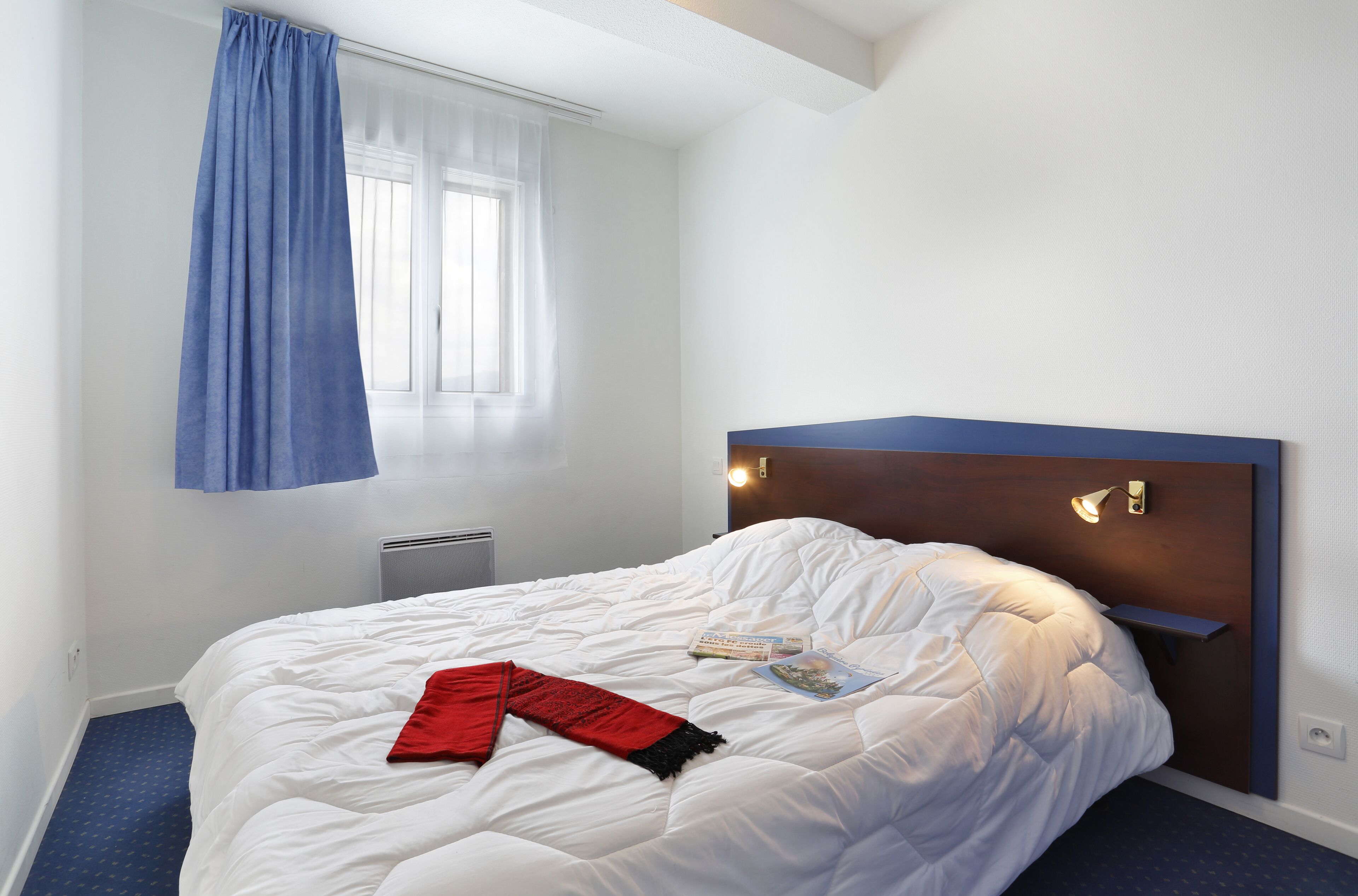 A neatly made bed with a white comforter and a red throw blanket, a bedside table with a lamp, and a window with a blue curtain.