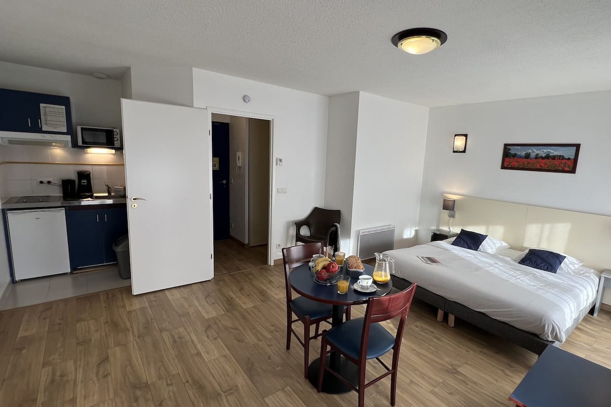 A compact hotel room with a bed, bedside table, and a small kitchenette.