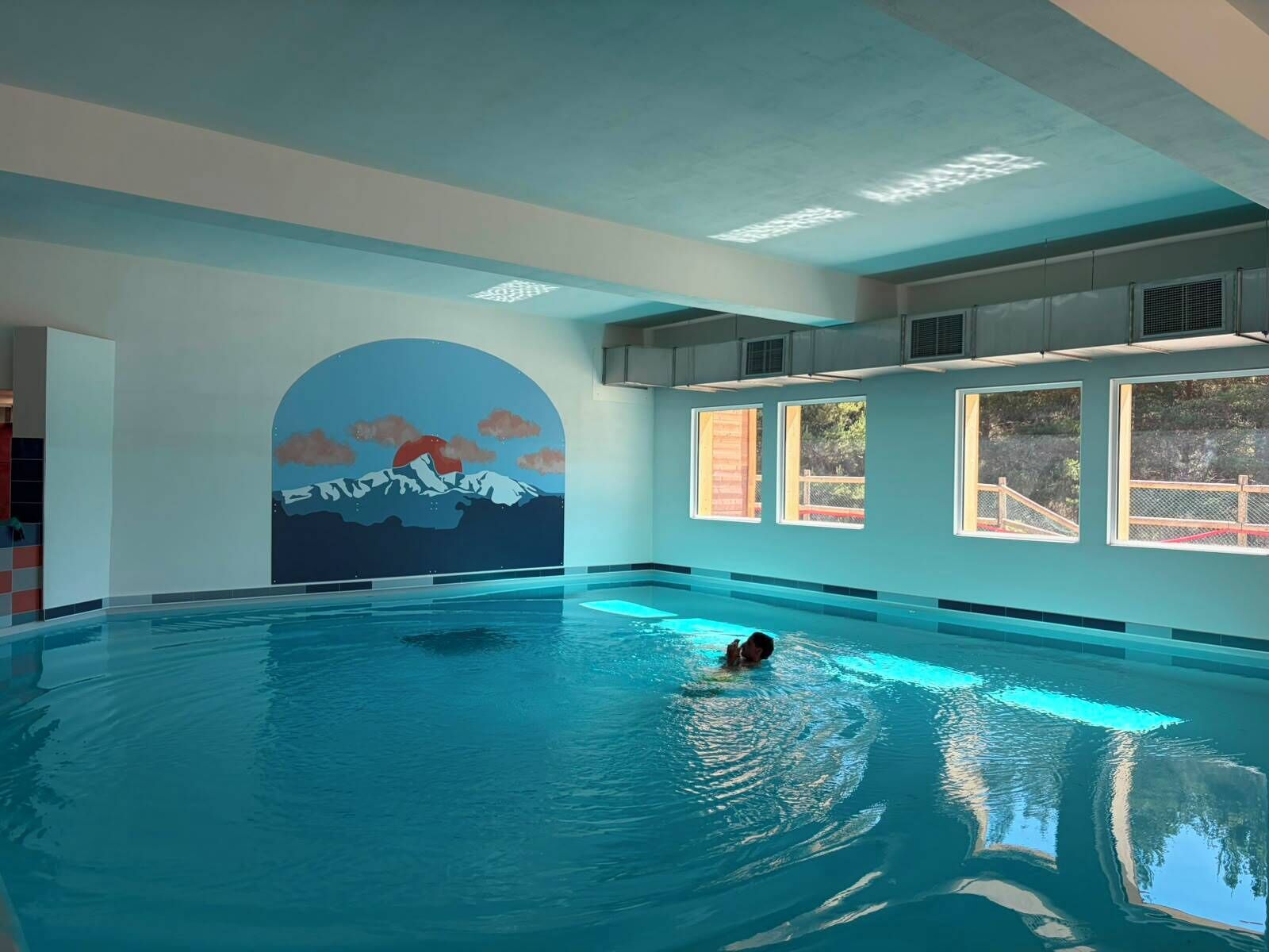 An indoor swimming pool with a mural of a mountain landscape, large windows, and a person swimming.