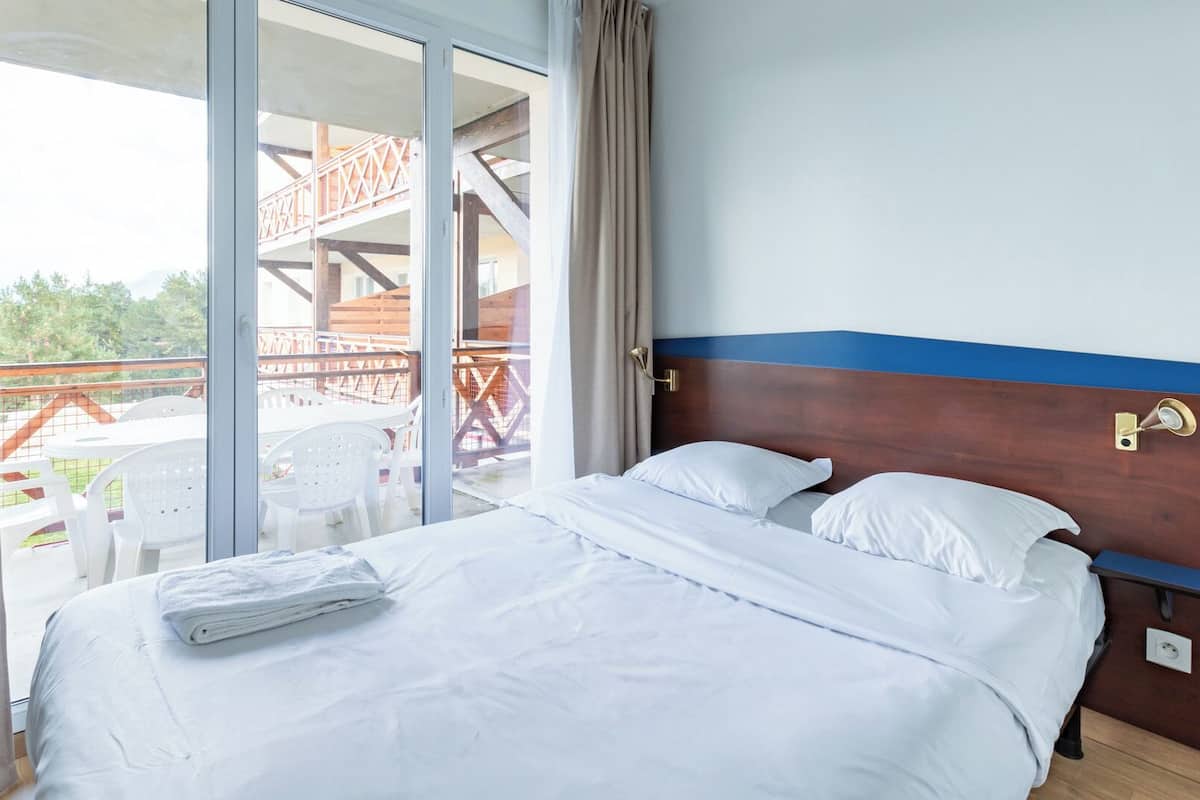A hotel room with a bed, a view of an outdoor balcony with chairs, and a wooden structure in the background.