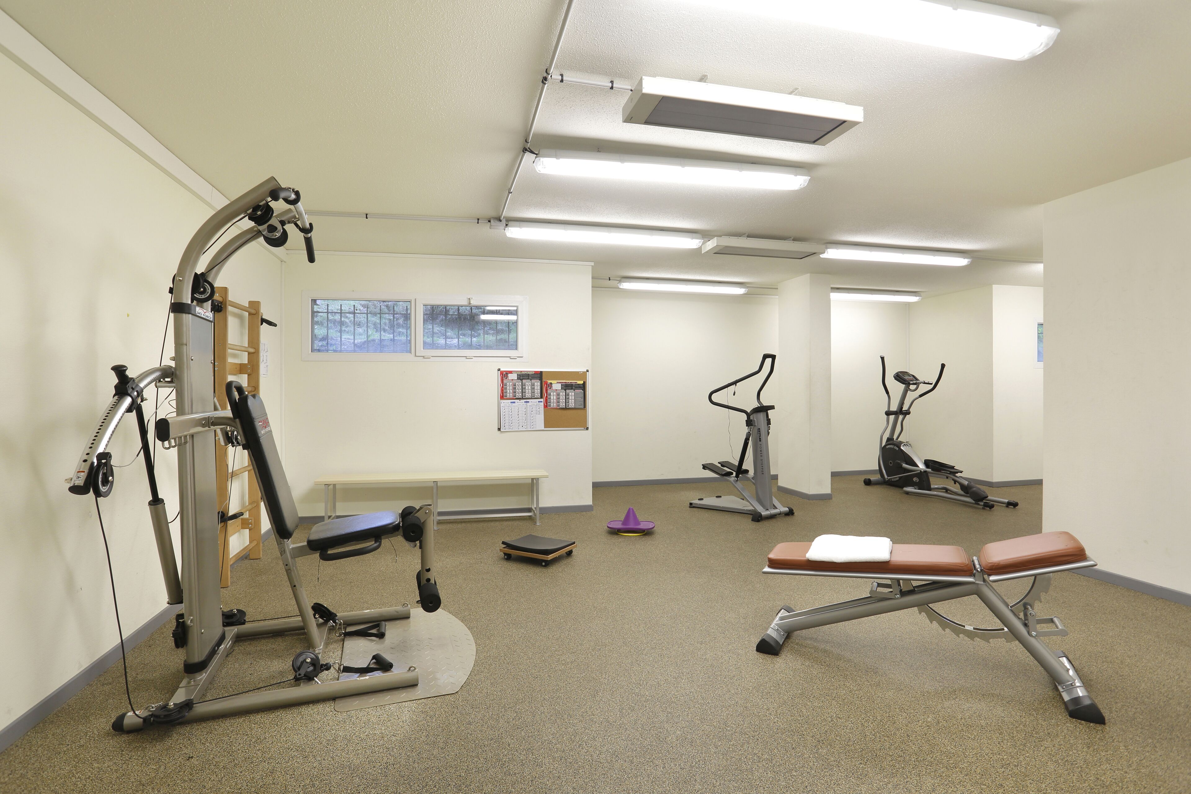 A gym with various exercise equipment, a bench, and a bulletin board on the wall.
